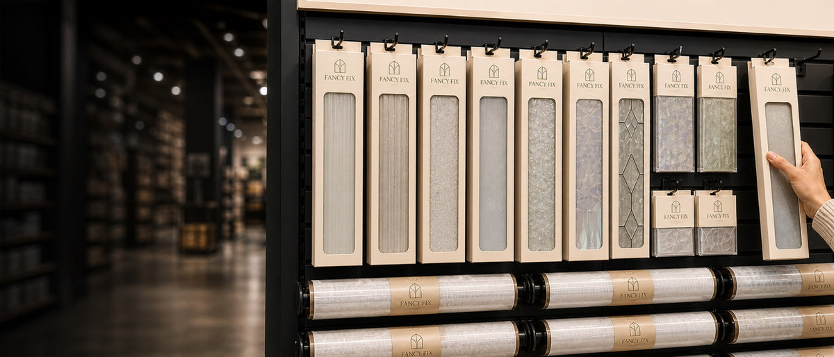 A hand lifting a decorative window film sample board from a retail display wall, with printed rolls stacked on the shelf below.