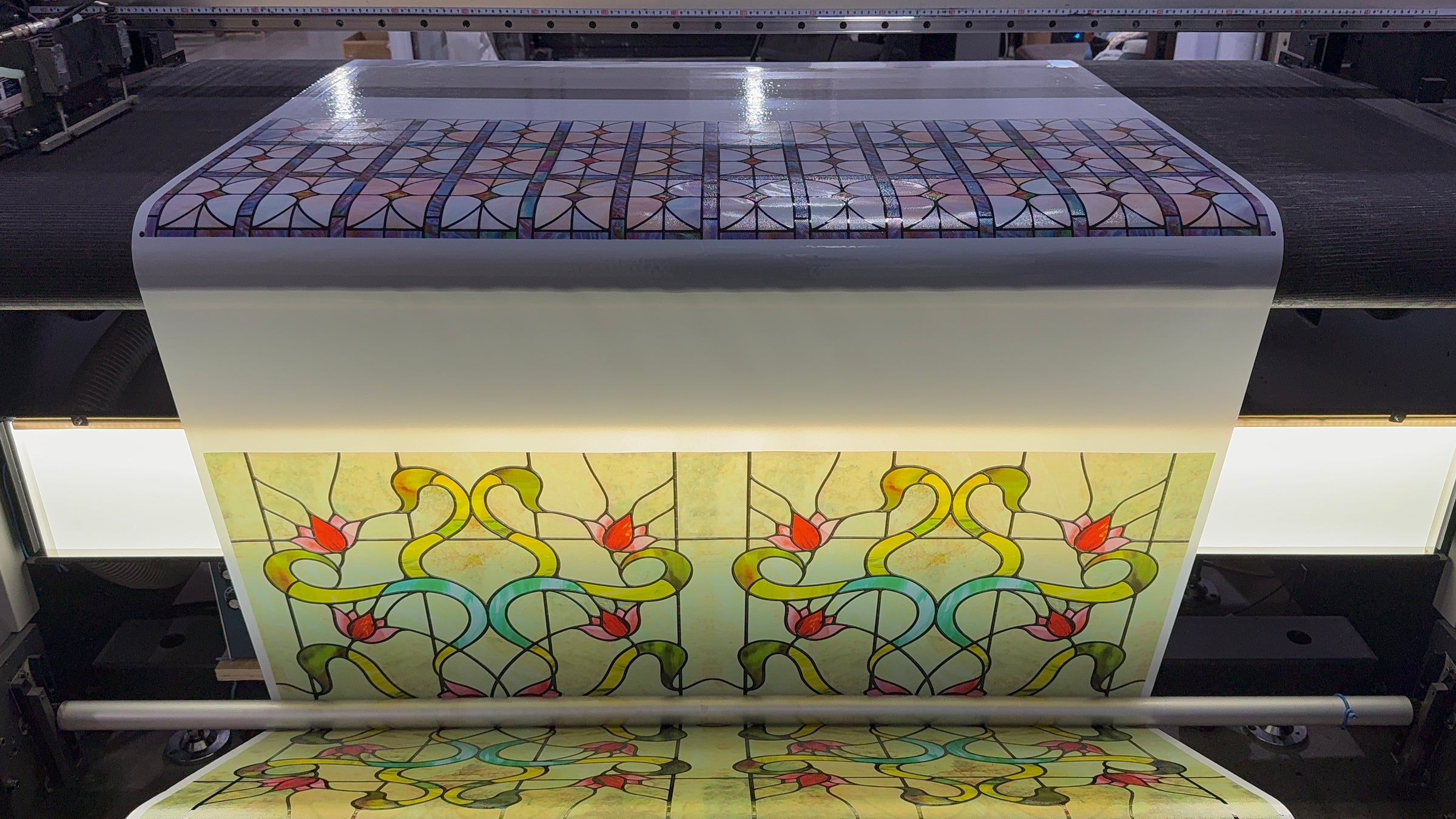 An original stained-glass pattern printing on a wide-format press at the FancyFix factory, backlit floral leaded-glass panels coming off the roll.
